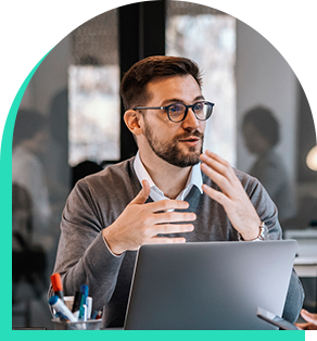 Picture of a bearded man with glasses gesturing to off-screen people with a laptop computer in front of him.